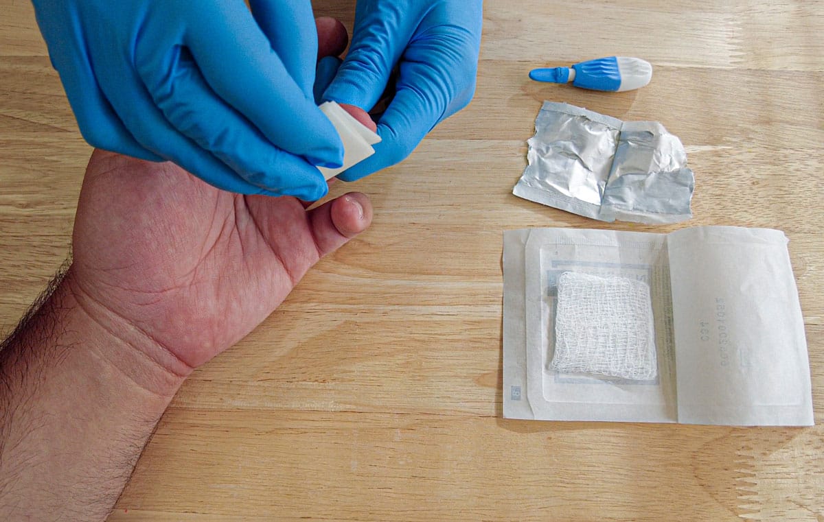 Gloved hands collecting fingerprick blood sample with prep pad and gauze